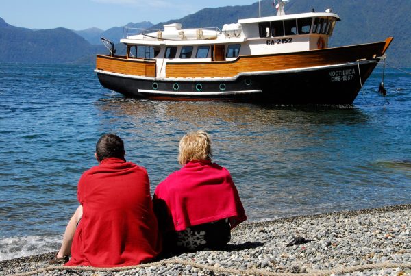 Two people sitting on a Chilean shoreline looking at a floating expedition yacht 