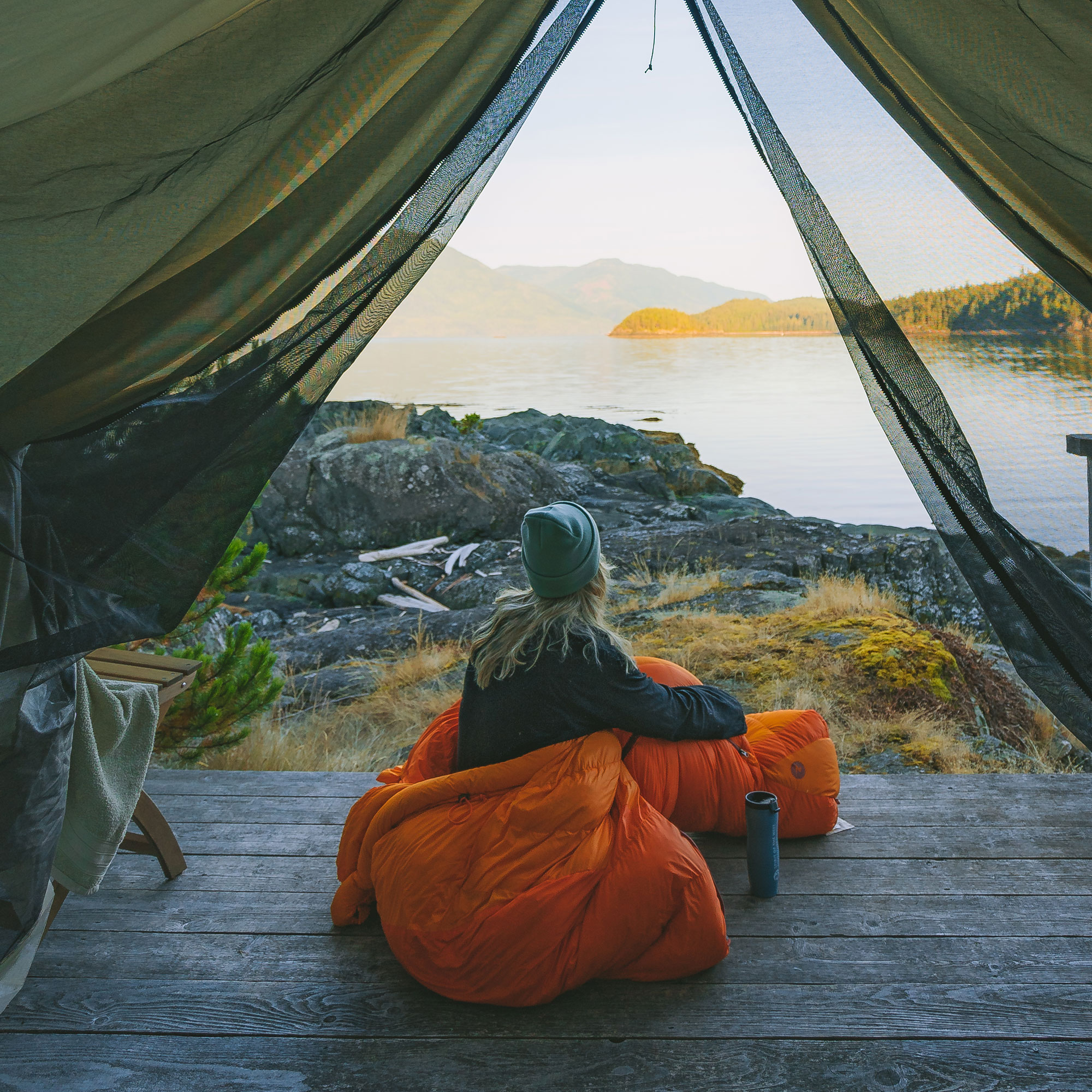 Person Wrapped in Orange Sleeping Bag in Open Tent Looking at Coastal BC Wilderness